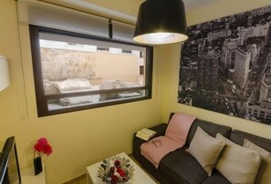 A room with a window, a sofa, a small table with a vase, and a cityscape painting on the wall. - Duque 2D · Standard Apartment without Terrace · 2 Bedrooms · 2 Bathrooms (Cordoba)