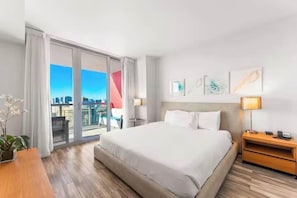 Ein Hotelzimmer mit einem großen Bett, einem Nachttisch aus Holz, einem Schreibtisch und Blick auf die Stadt durch ein großes Fenster.