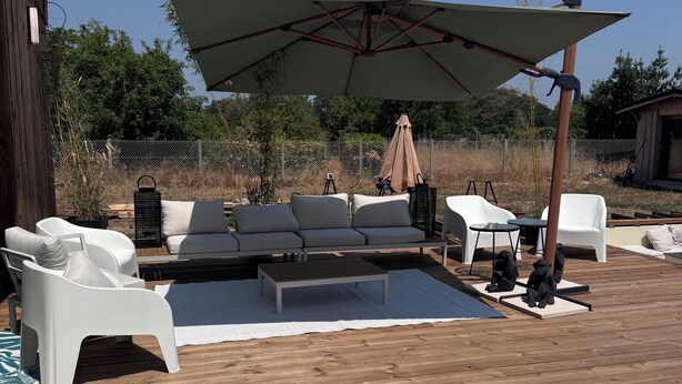 Een terras buiten met een houten terras, witte meubels en een grote parasol.