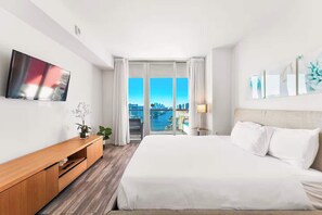 A hotel room with a bed, a TV, a desk, a lamp, and a view of a cityscape. - Bright 1BR - Sunset View -Beach Access (Hallandale Beach)