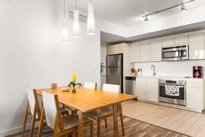 A modern kitchen with stainless steel appliances, a wooden dining table, and pendant lighting. - Bright 1BR - Sunset View -Beach Access (Hallandale Beach)