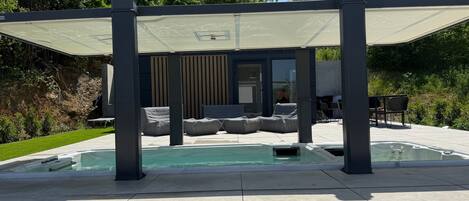 Un área de piscina moderna al aire libre, con zona de estar cubierta y un jacuzzi.