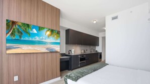 A modern bedroom with a large bed, a kitchen area with dark cabinets, and a beach-themed painting on the wall.