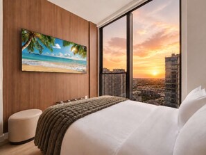 Ein Schlafzimmer mit großem Fenster, einem Bett, einem am Wand montierten Fernseher und Blick auf eine Skyline bei Sonnenuntergang.