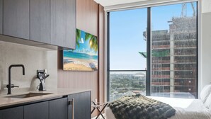 A modern bedroom with a large window, a bed with a blanket, a painting of a beach, and a view of a construction site. - Stunning Studio Suite At The E11EVEN Residences (Miami)