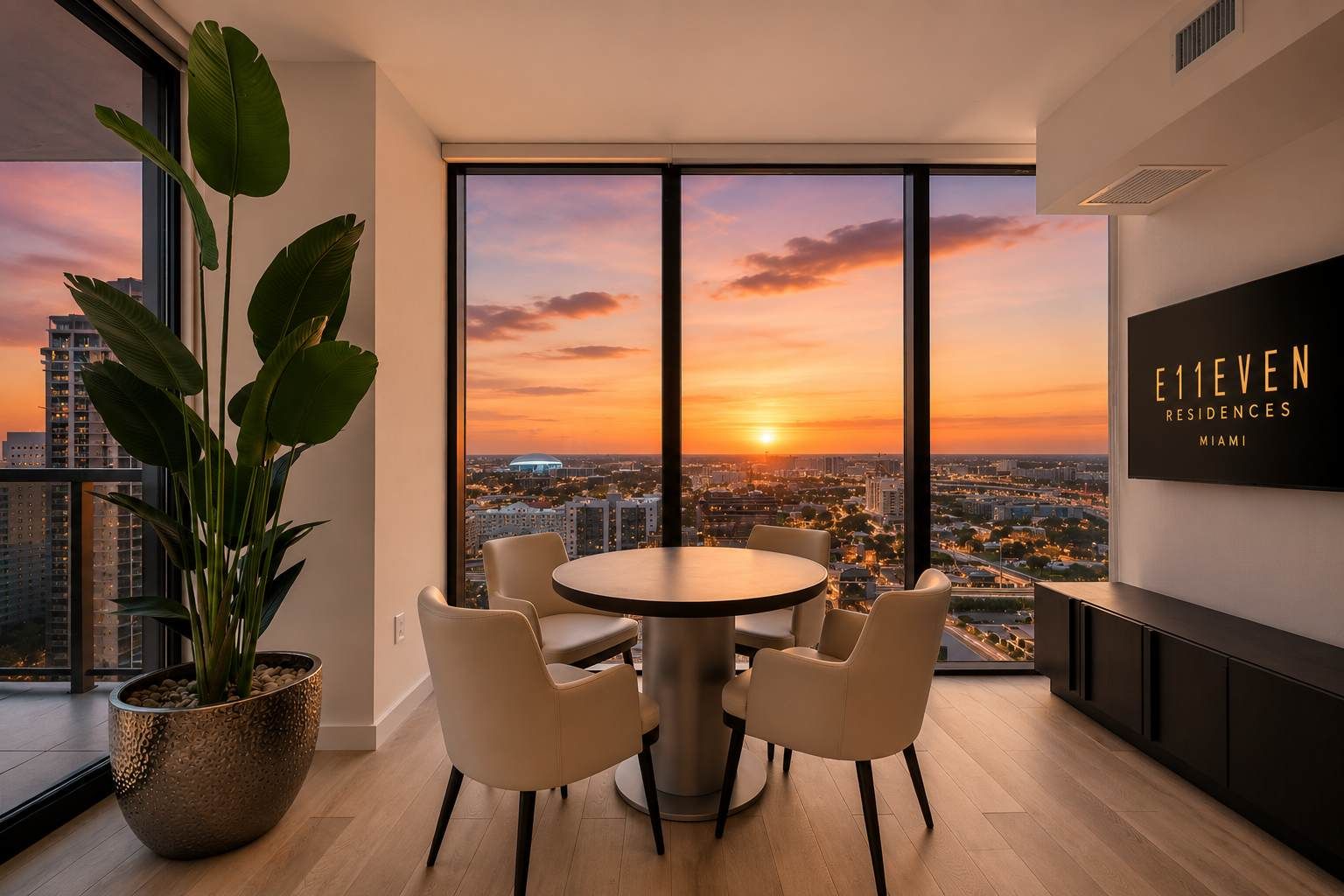 A modern dining area with a round table and chairs, large windows with a sunset view, and a potted plant.