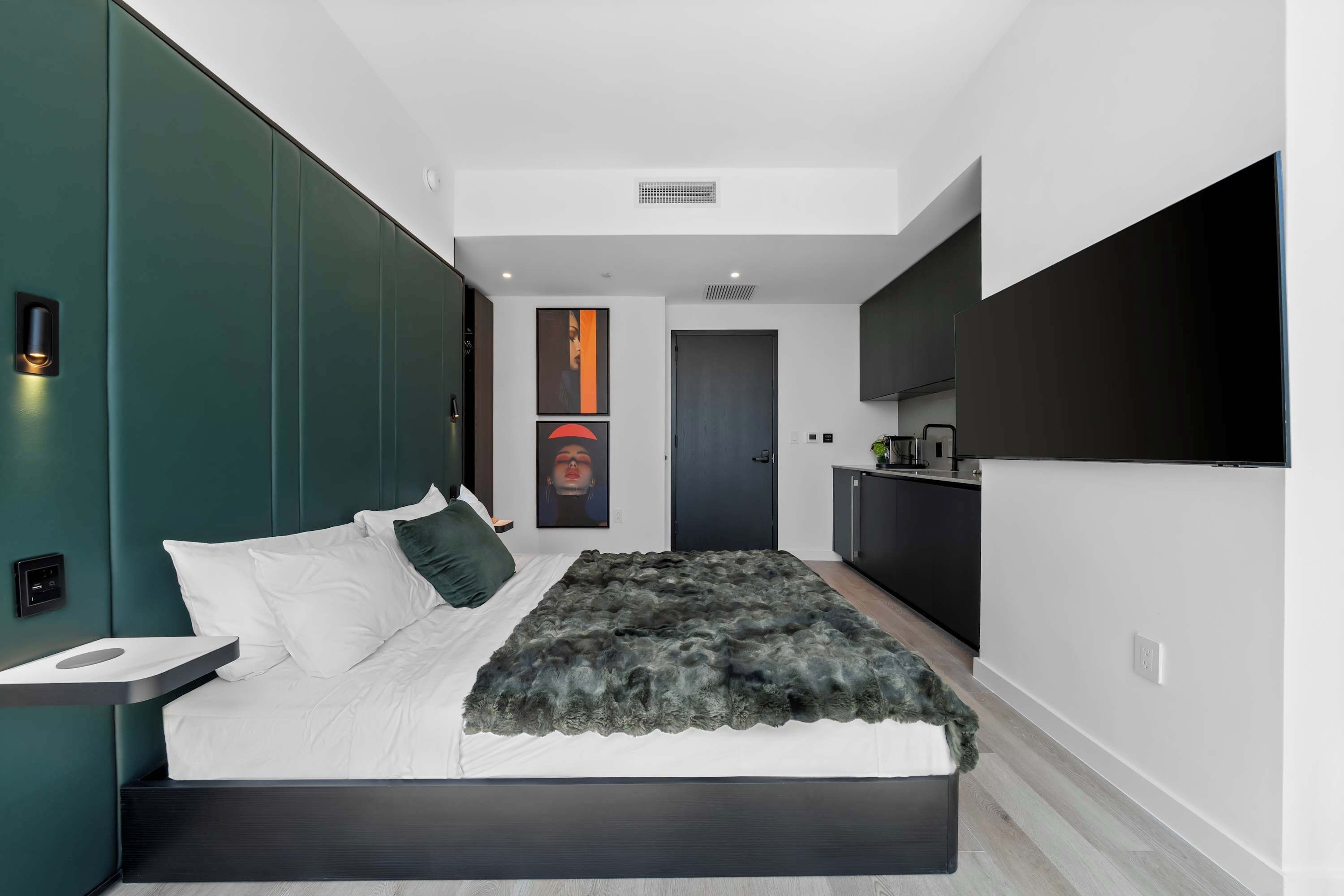 Een moderne slaapkamer met een bed, nachtkastje, kunstwerk en uitzicht op de keuken.