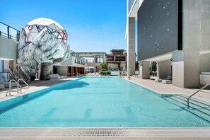 Ein Schwimmbecken mit einer großen Handskulptur, moderne Gebäude und ein klarer Himmel.