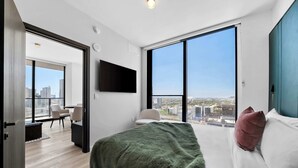 Ein modernes Hotelzimmer mit einem großen Bett, einem an der Wand befestigten Fernseher und Blick auf die Stadt durch große Fenster.
