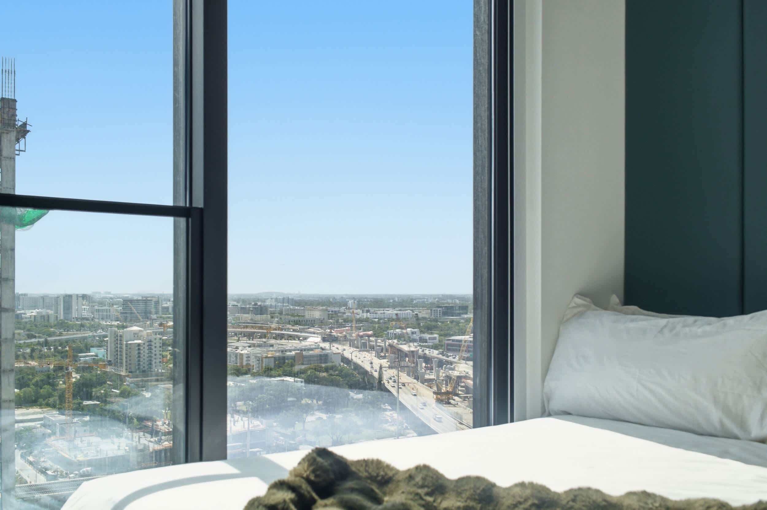 Un dormitorio con un ventanal que ofrece vistas a la ciudad, una cama con ropa blanca y vistas a una obra en construcción.