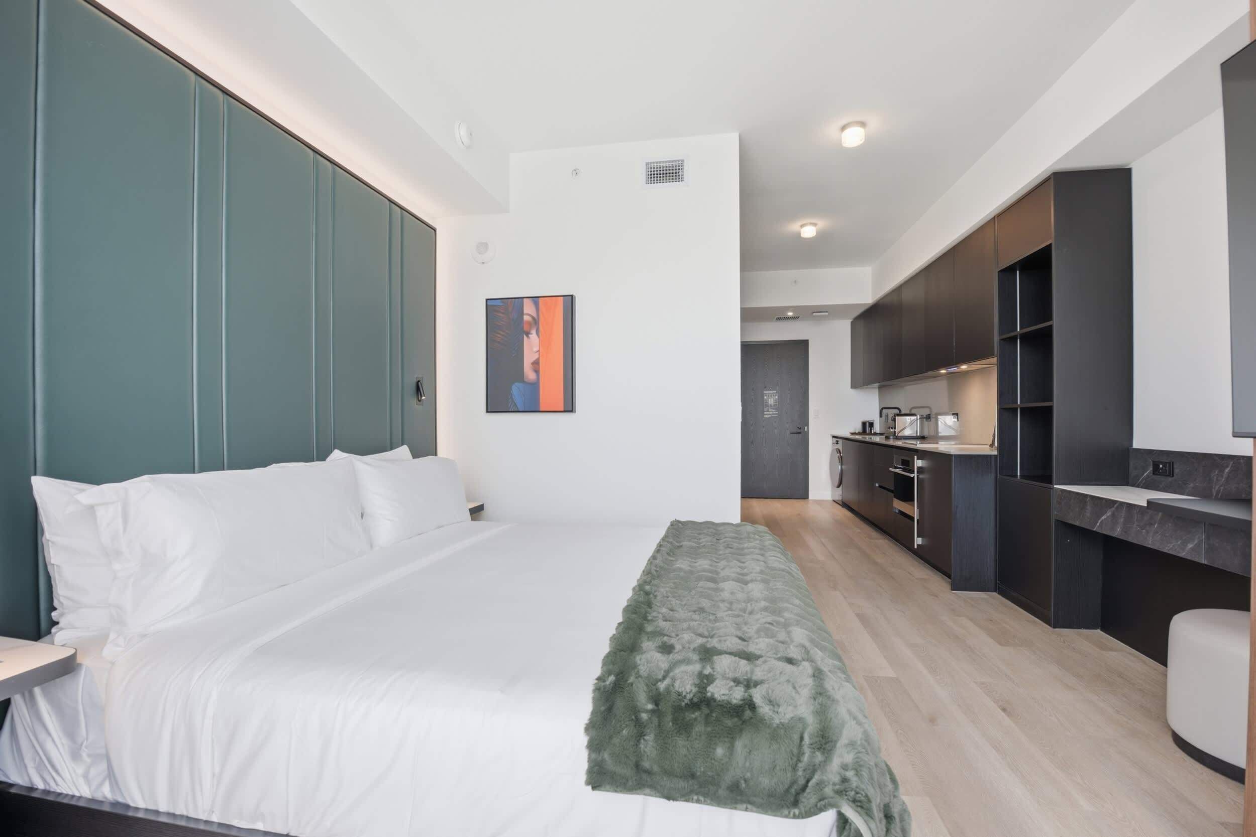 A modern bedroom with a large bed, a bench, and a kitchenette in the background.