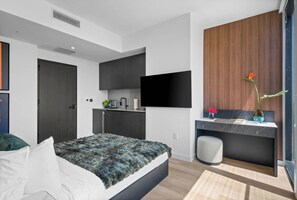 Ein modernes Schlafzimmer mit Bett, Schreibtisch, Fernseher und einer kleinen Küchenzeile.