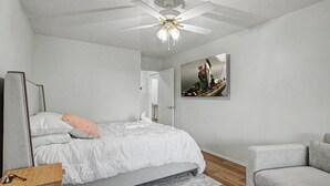 Een slaapkamer met een bed, een grijze bank, een plafondventilator, een schilderij en een nachtkastje van hout.