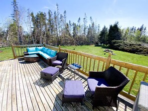 Eine Holzterrasse mit Gartenmöbeln, darunter eine Sitzecke und Stühle, mit Blick auf eine Rasenfläche mit Bäumen.