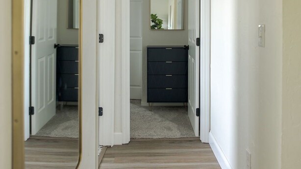 A hallway with a wooden floor, white walls, a mirror, a vent, and a dark-colored dresser. - The Village 3BR (The Village)
