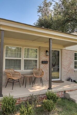A covered porch with two wicker chairs and a small table, a brick wall, and a clear sky. - The Village 3BR (The Village)