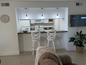 Eine moderne Küche mit weißen Schränken, einem eingebauten Backofen und einer Mikrowelle. Auf der Arbeitsplatte stehen zwei weiße Barhocker und eine grüne Tasse. An der Wand hängt eine Uhr, und über der Arbeitsplatte ist ein Fernseher angebracht.