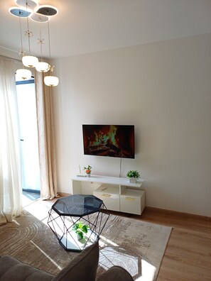 Un saló modern amb televisió de pantalla plana penjada a la paret, una taula de centre de vidre amb una planta, un moble baix blanc amb objectes decoratius i una finestra gran amb cortines.