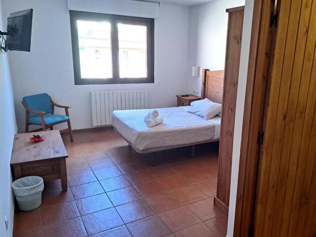 Un dormitorio con cama, una silla, una mesita, un radiador y una ventana.