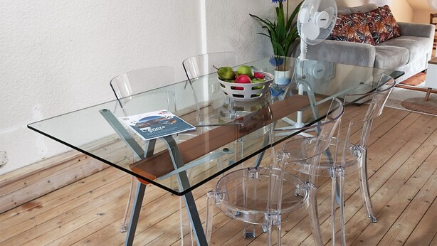 Un comedor moderno con una mesa de vidrio, sillas transparentes, suelo de madera y una pared blanca con obras de arte.