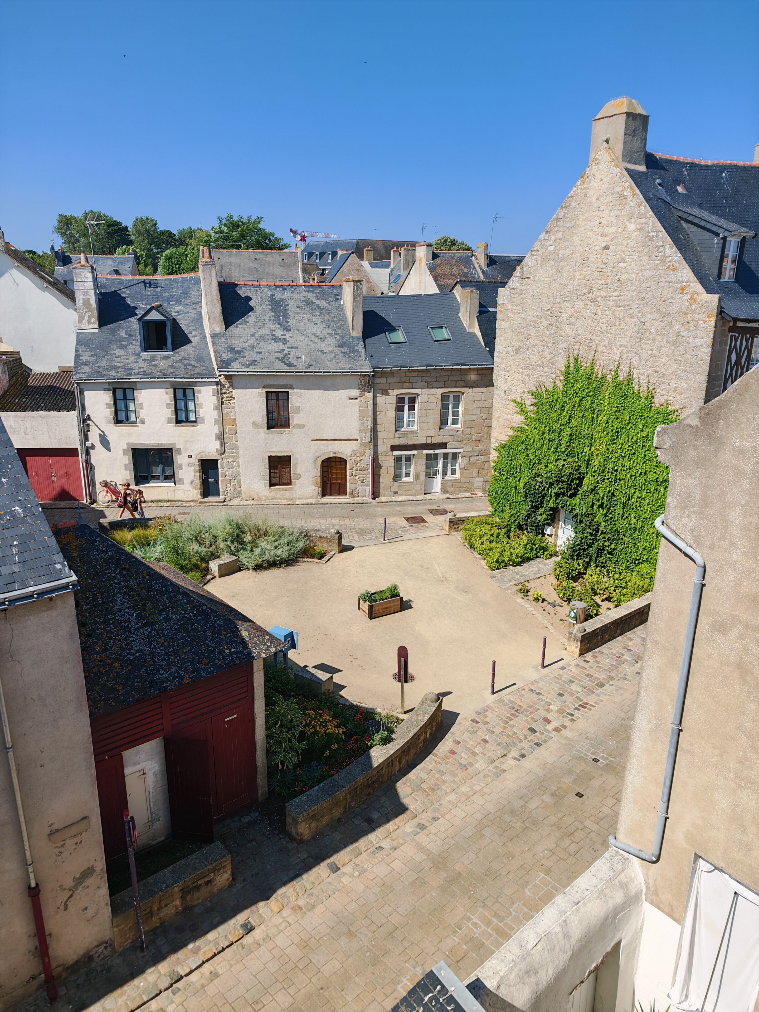 Une cour bordée de bâtiments en pierre, avec une porte rouge et un petit jardin.