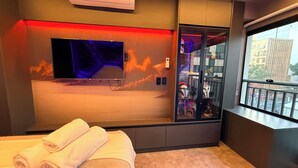Une chambre d’hôtel moderne équipée d’un téléviseur à écran plat, d’une vitrine contenant des objets de collection, d’une grande fenêtre donnant sur la ville et d’une serviette soigneusement pliée posée sur le lit.