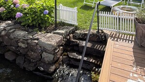 Una casa de dos pisos con un porche delantero, un muro de contención de piedra y una terraza de madera con sillas.