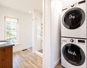 A laundry room with two stacked front-loading washers, a glass shower enclosure, and a window with a view of greenery. - DIRECT LAKEFRONT Property on Winsted Lake (Winchester)