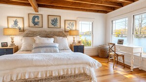 Un dormitorio con cama, mesitas de noche, lámparas, una obra de arte enmarcada, una silla y vistas a un lago a través de amplios ventanales.