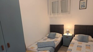 Un dormitorio pequeño y bien iluminado con dos camas individuales, una mesita de noche con lámpara y un armario.