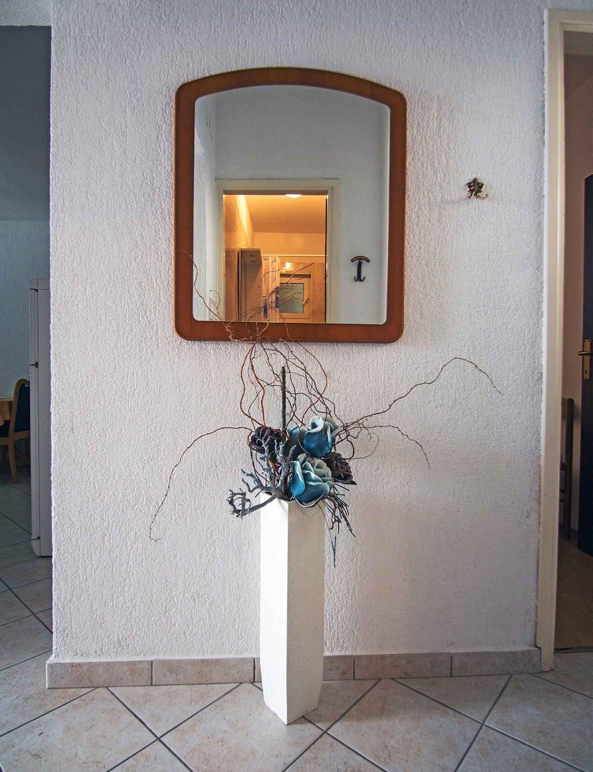 Un baño con pared blanca texturizada, un espejo con marco de madera y un lavamanos pedestal blanco con un arreglo decorativo de plantas.