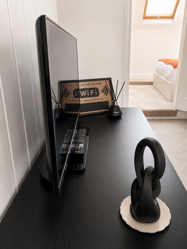A modern desk with a computer monitor, keyboard, and headphones. A sign with 'WiFi' and a password prompt is visible.