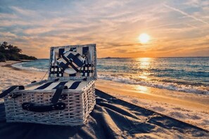 A wicker basket with a black handle on a beach with the ocean and a sunset in the background.