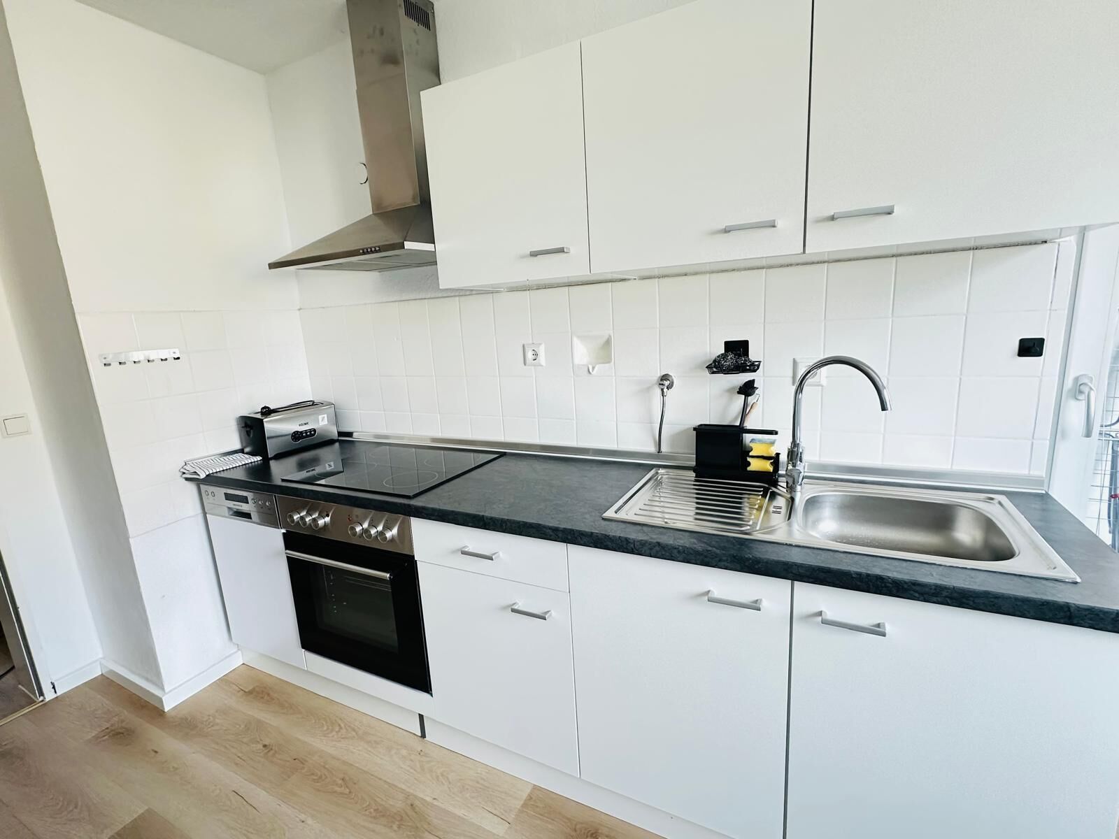 Une cuisine moderne avec des meubles blancs, un évier en acier inoxydable, un four encastré et une plaque de cuisson noire.
