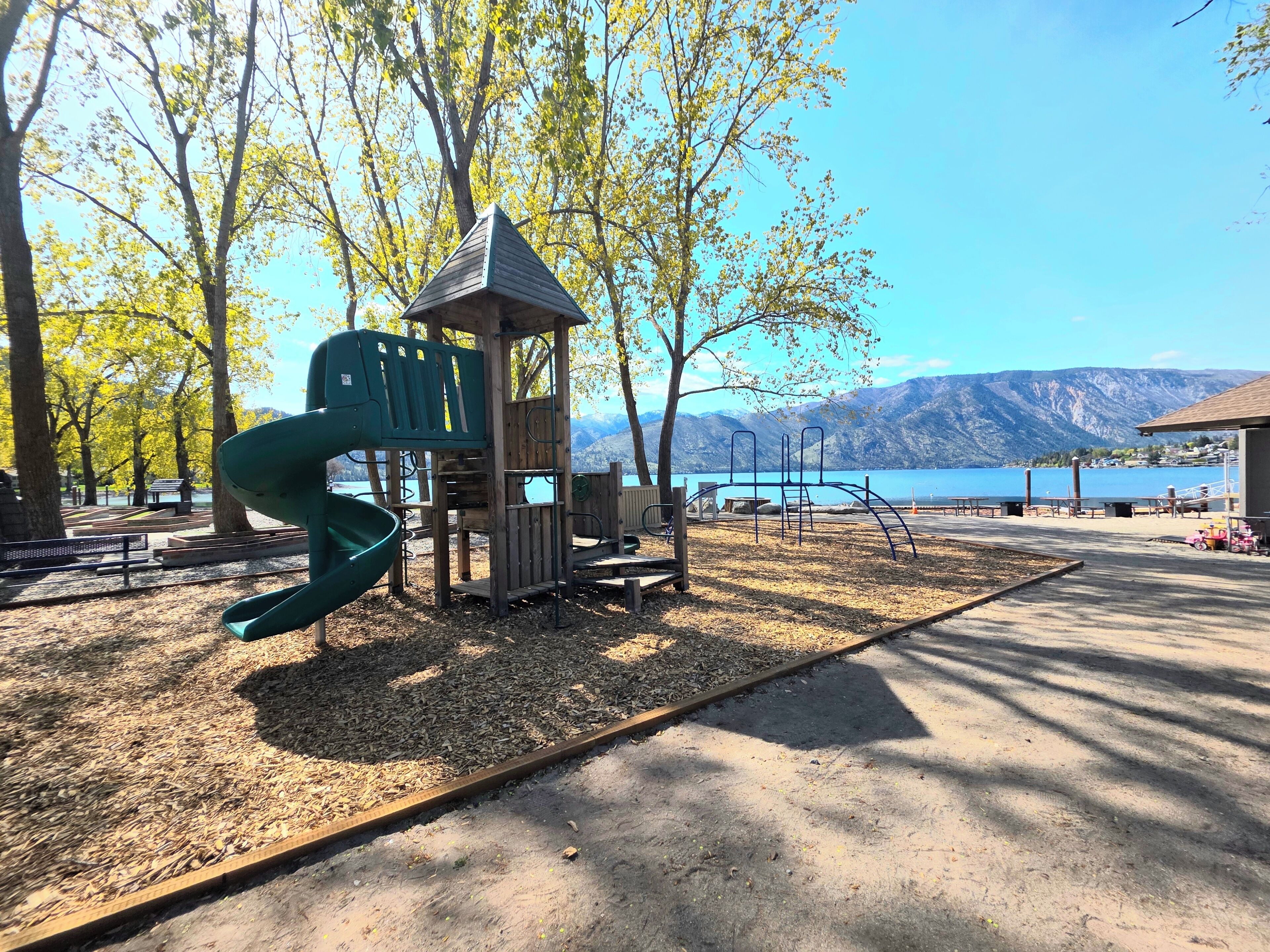 A playground with a slide and climbing structures, benches, and a view of a lake with mountains in the background.