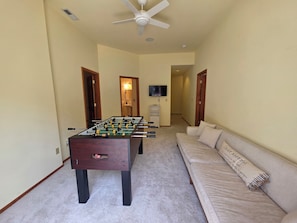 A room with a foosball table, a sofa, a television, and a ceiling fan.