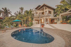 Ein zweistöckiges Haus mit Pool, umgeben von Palmen und einer überdachten Terrasse.