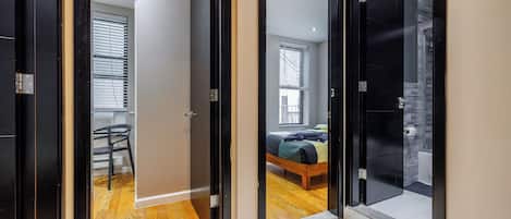 A hallway with a bed, a chair, and a window in the adjacent room.