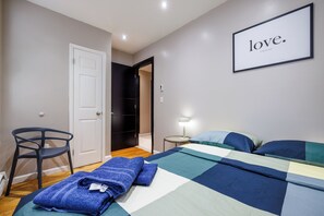 Ein Schlafzimmer mit einem Bett, einem Sessel, einem Nachttisch mit Lampe und einer Tür zu einem weiteren Raum.