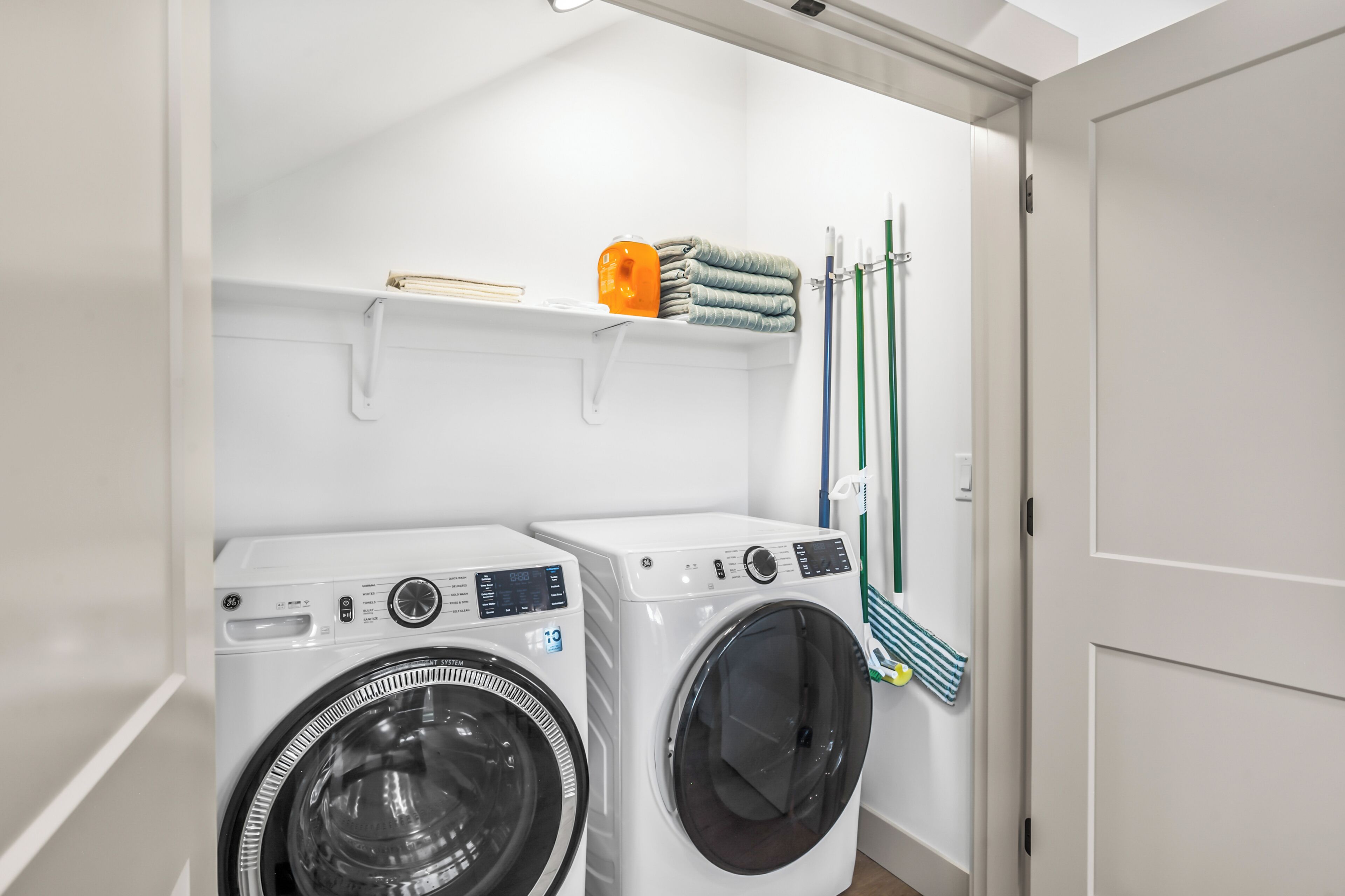 A laundry area with two front-loading washing machines, a shelf with folded towels, and a rack with cleaning supplies.
