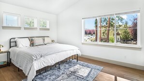 A bedroom with a bed, a window with a view of trees, and a bedside table with a lamp. - NEW Downtown Sisters Luxe Retreat with Hot Tub + Firepit (Sisters)