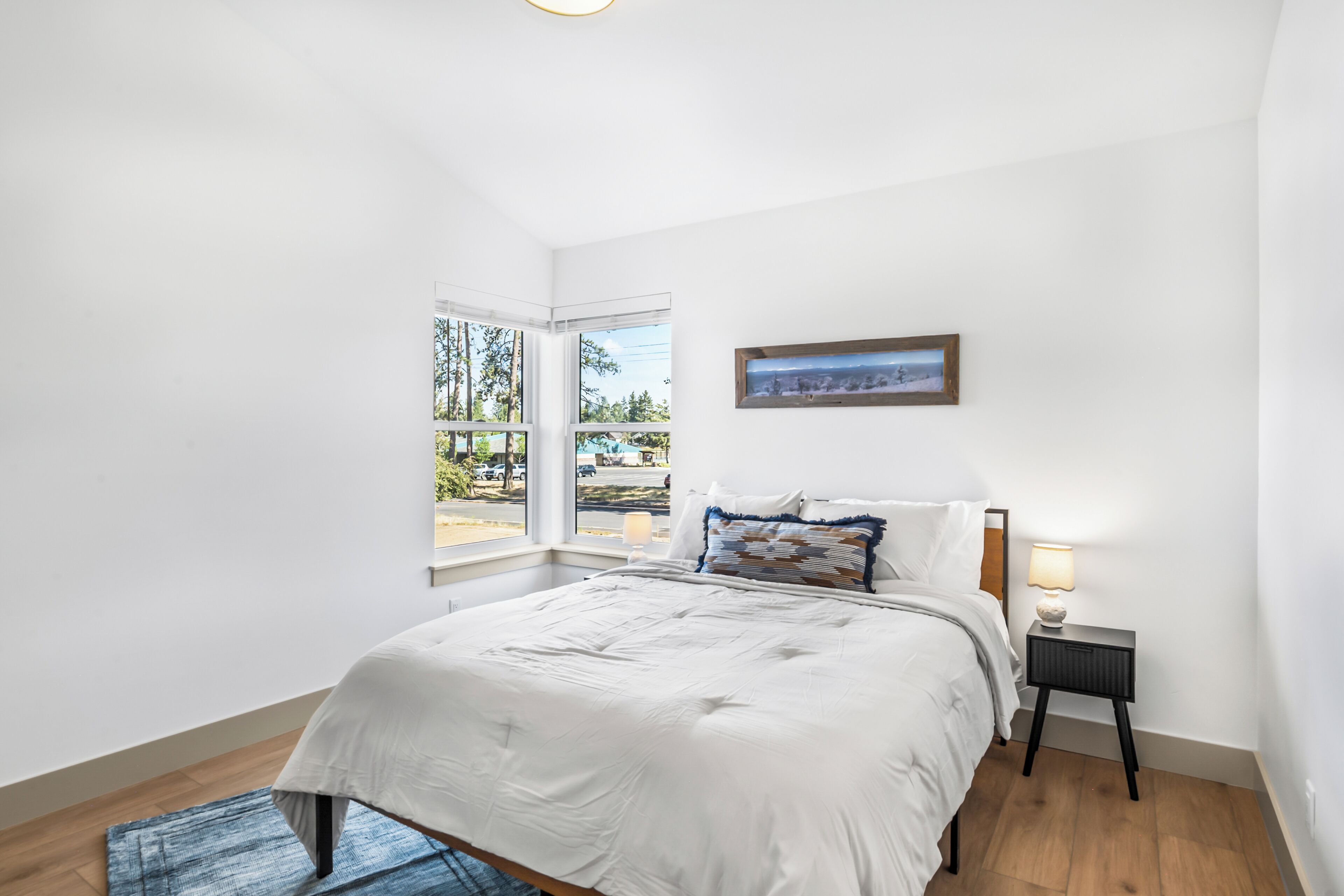 Ein Schlafzimmer mit Bett, Nachttisch, Lampe, Fenster mit Aussicht und einem gerahmten Bild an der Wand.