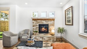 A living room with a fireplace, a grey armchair, a coffee table with books and decorative items, and a plant. - NEW Downtown Sisters Luxe Retreat with Hot Tub + Firepit (Sisters)