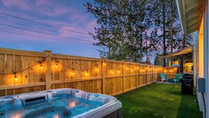 Un patio trasero con jacuzzi, cerca de madera, luces colgantes y césped.