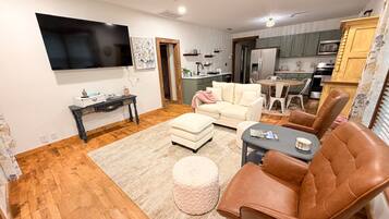 Una sala de estar con sofá, reposapiés y sillones, un comedor con mesa y sillas, y una cocina con alacenas y electrodomésticos.