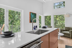 A modern kitchen with a large window, a sink, and a dining area with a green sofa. - New Home + Guest House on Bike Trail (Bella Vista)