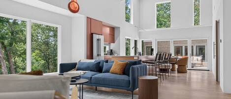 A spacious living area with a sofa, coffee table, and dining table. There is a kitchen in the background with a refrigerator and cabinets. Large windows provide natural light and views of the outdoors.