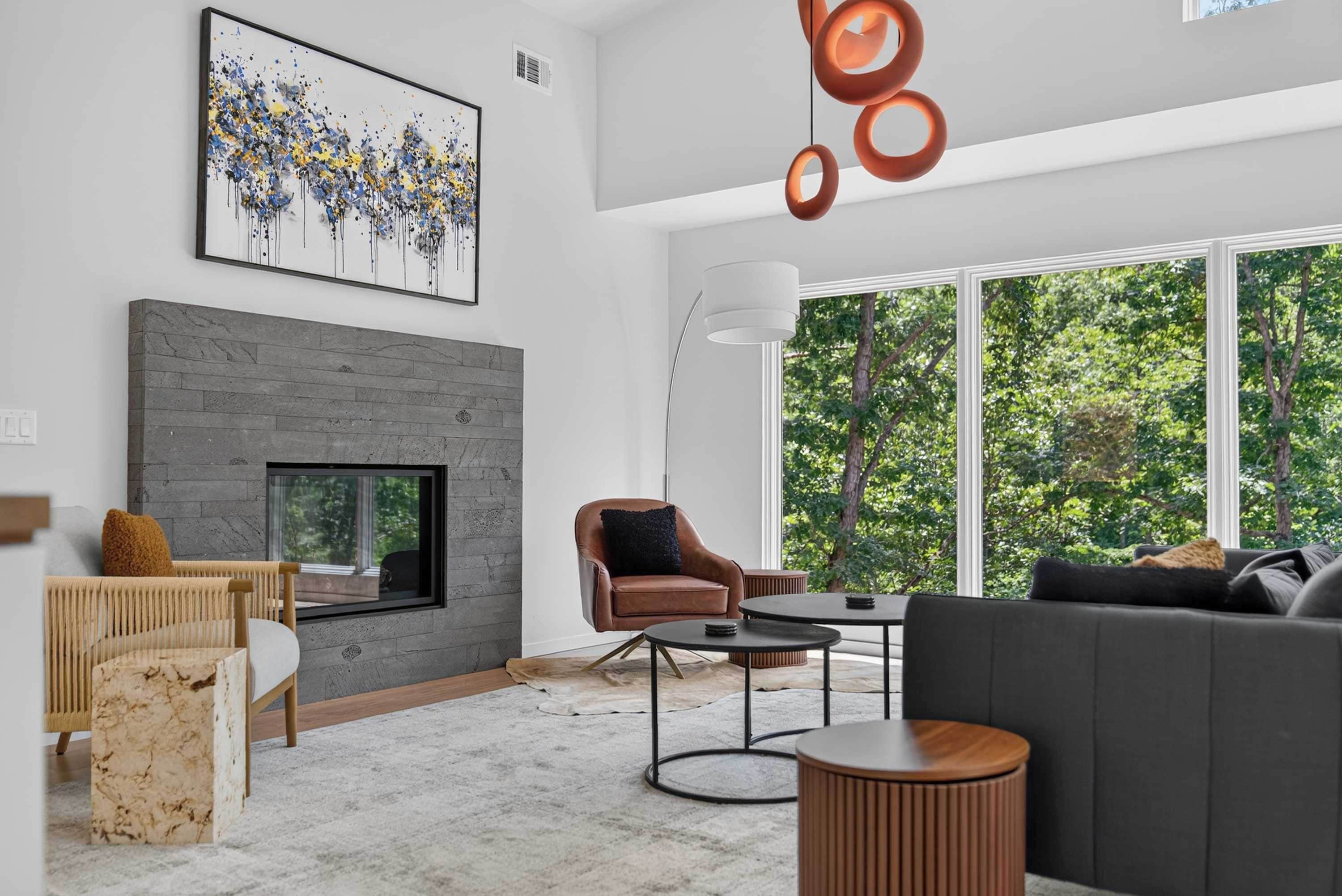 A modern living room with a fireplace, a sofa, a coffee table, and a large window with a view of trees.