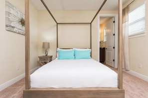 A four-poster bed with white bedding and two light blue pillows, a wooden nightstand with a lamp, and a wall-mounted painting. - Luxury Dreams Disney Home Private Pool Spa Gameroom (Kissimmee)