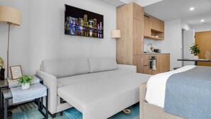 Quarto de hotel moderno com sofá, cama, escrivaninha com luminária, pintura de paisagem urbana e cozinha compacta.
